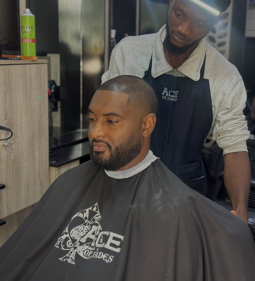 A barber in an Ace of Fades apron finishing a client's low fade and beard